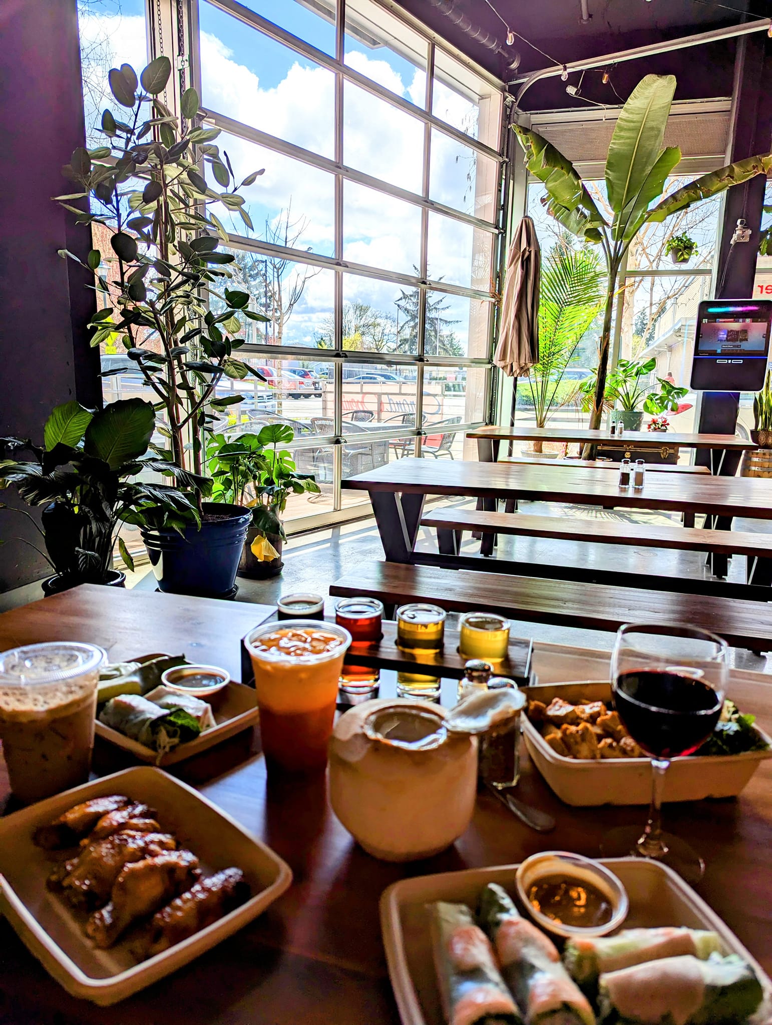 Variety of Vietnamese dishes and craft beverages at La Ca Cafe Tacoma including summer rolls, appetizers, and colorful drinks on communal wooden table with large windows, natural light, and tropical plants creating welcoming atmosphere
